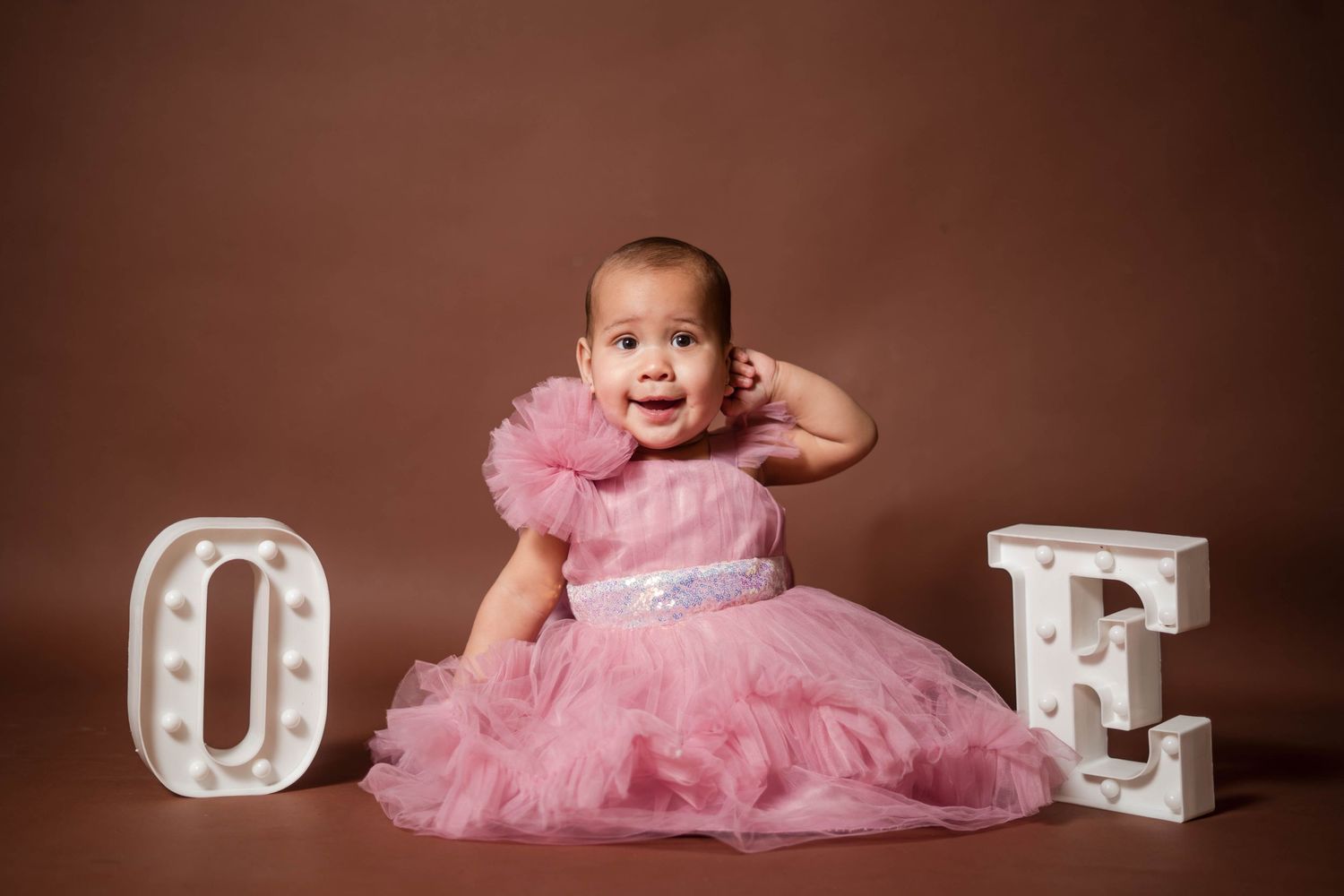 Happy baby girl in pink tulle dress posing between glowing letter O and E marquee signs.
