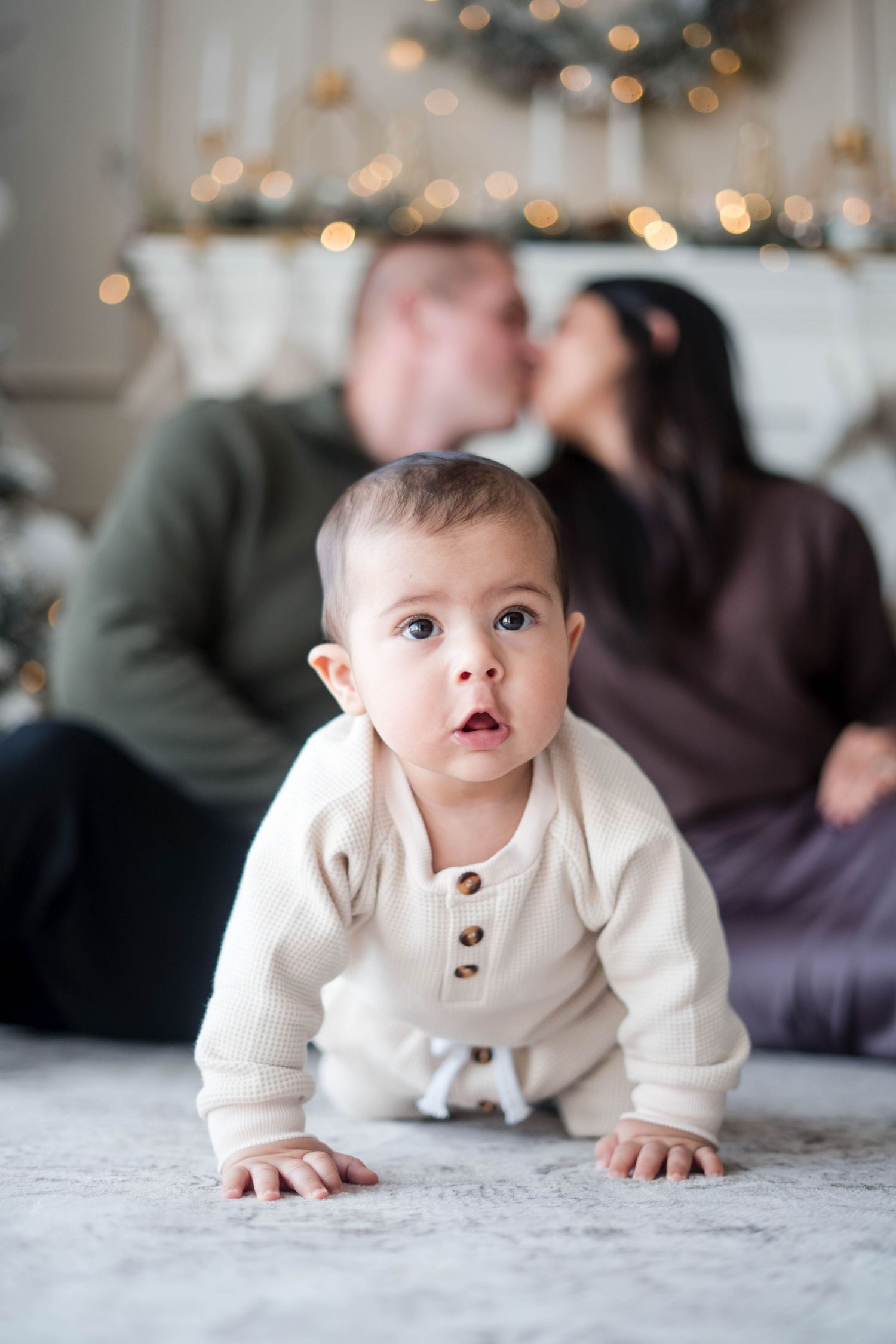 Curious baby in cream onesie crawls on floor while parents kiss near Christmas tree in background.