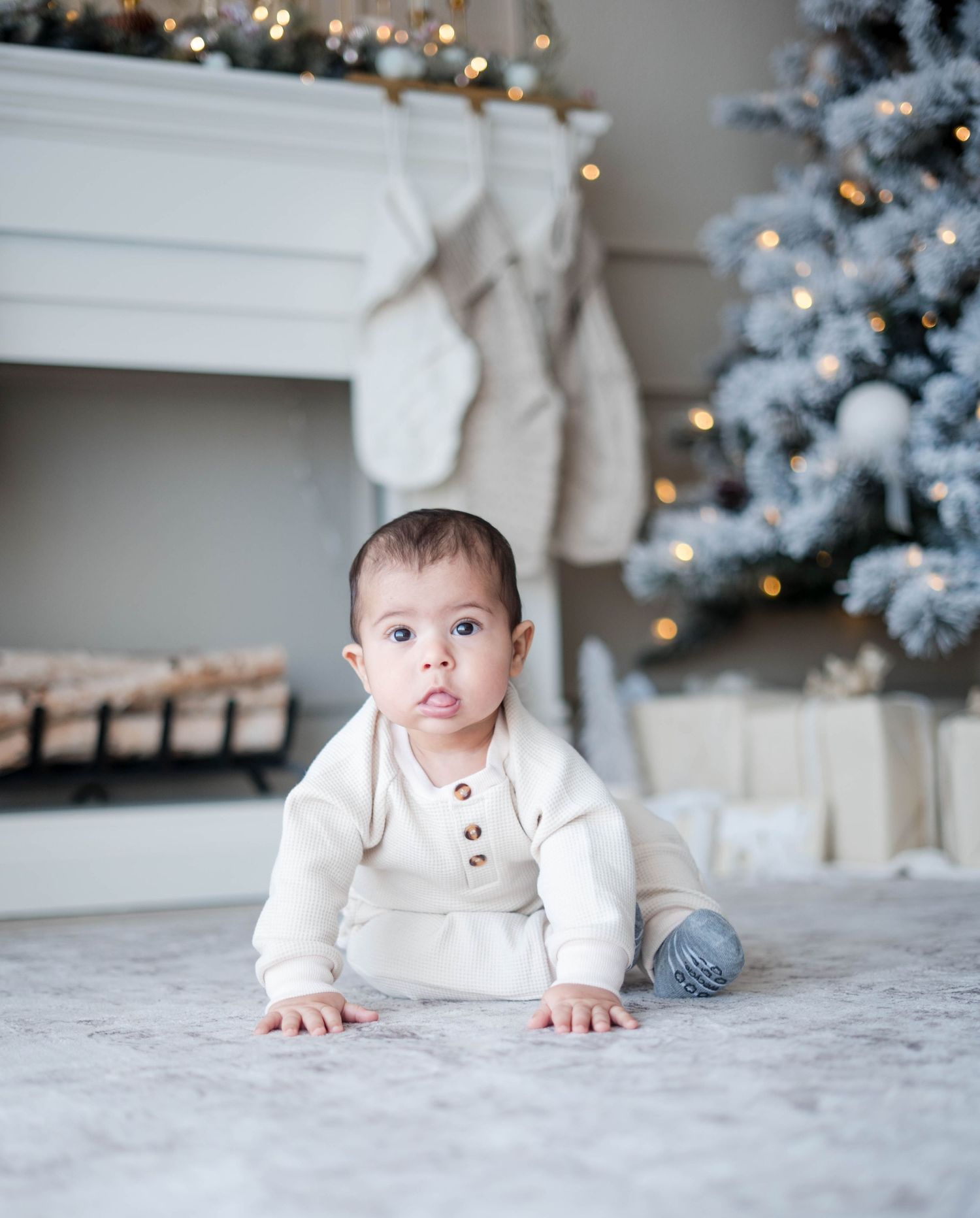 Adorable baby in cream button onesie crawls on floor near decorated Christmas tree and fireplace.