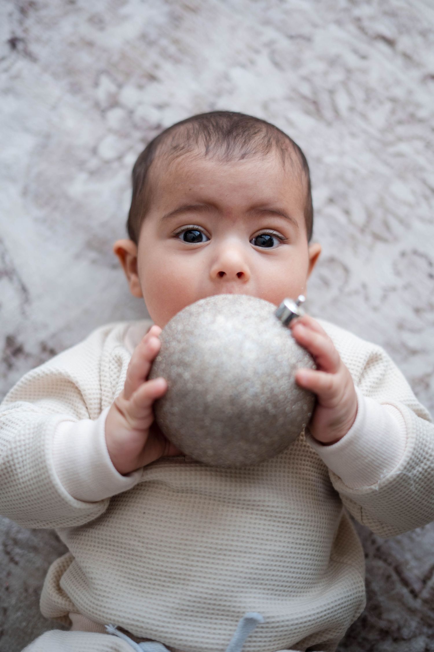 Curious baby in white outfit holding a silver glitter Christmas ornament ball up to their mouth.