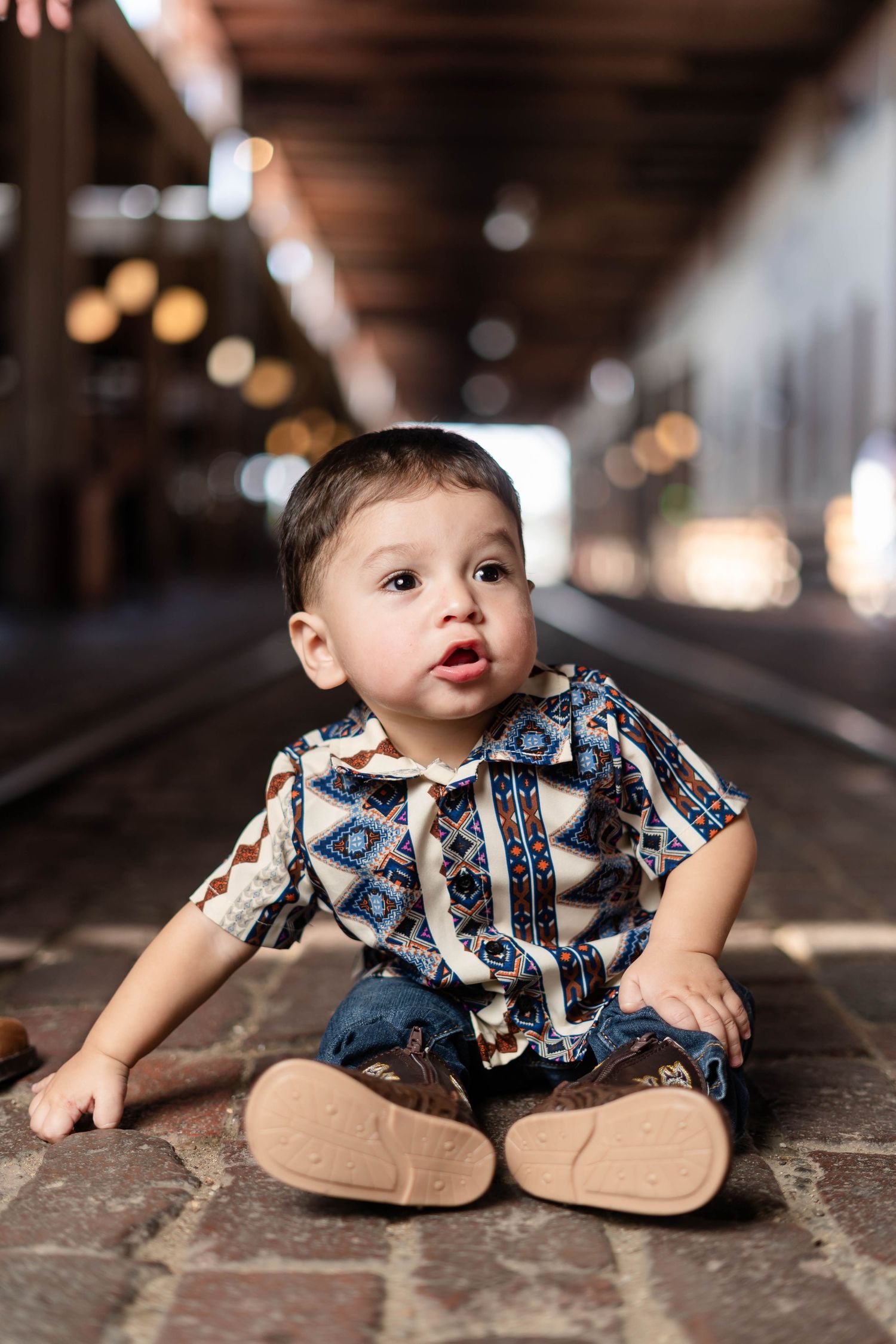 Cute baby boy in patterned shirt and jeans sitting on cobblestone floor indoors.