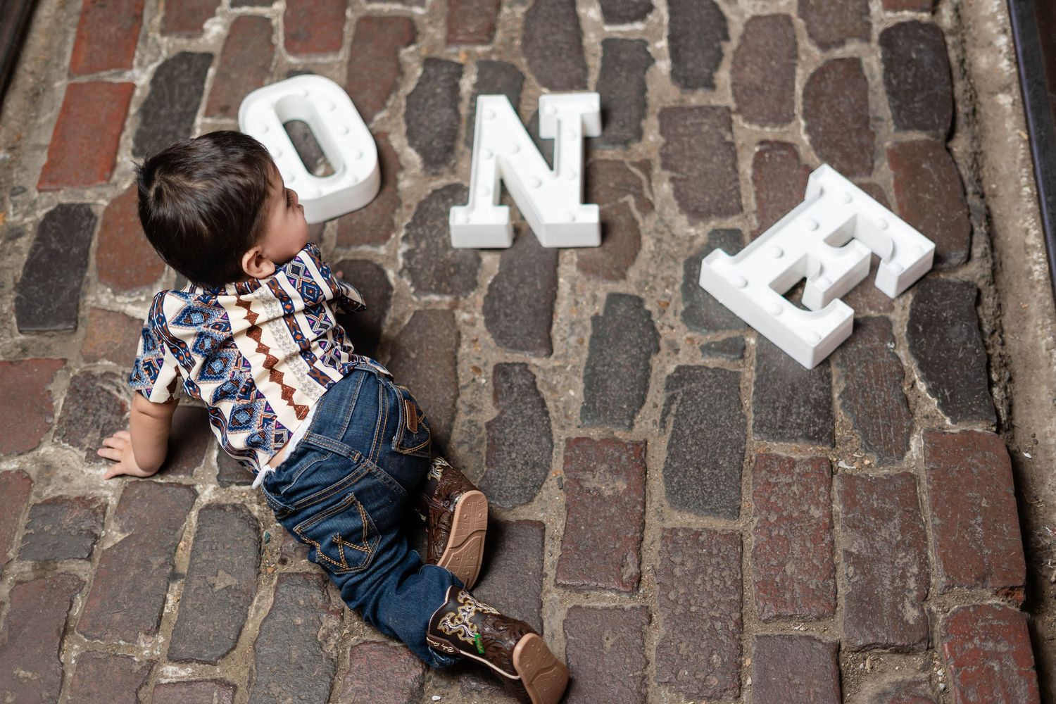 Overhead view of baby boy sitting on cobblestone pavement next to white ONE letter props.