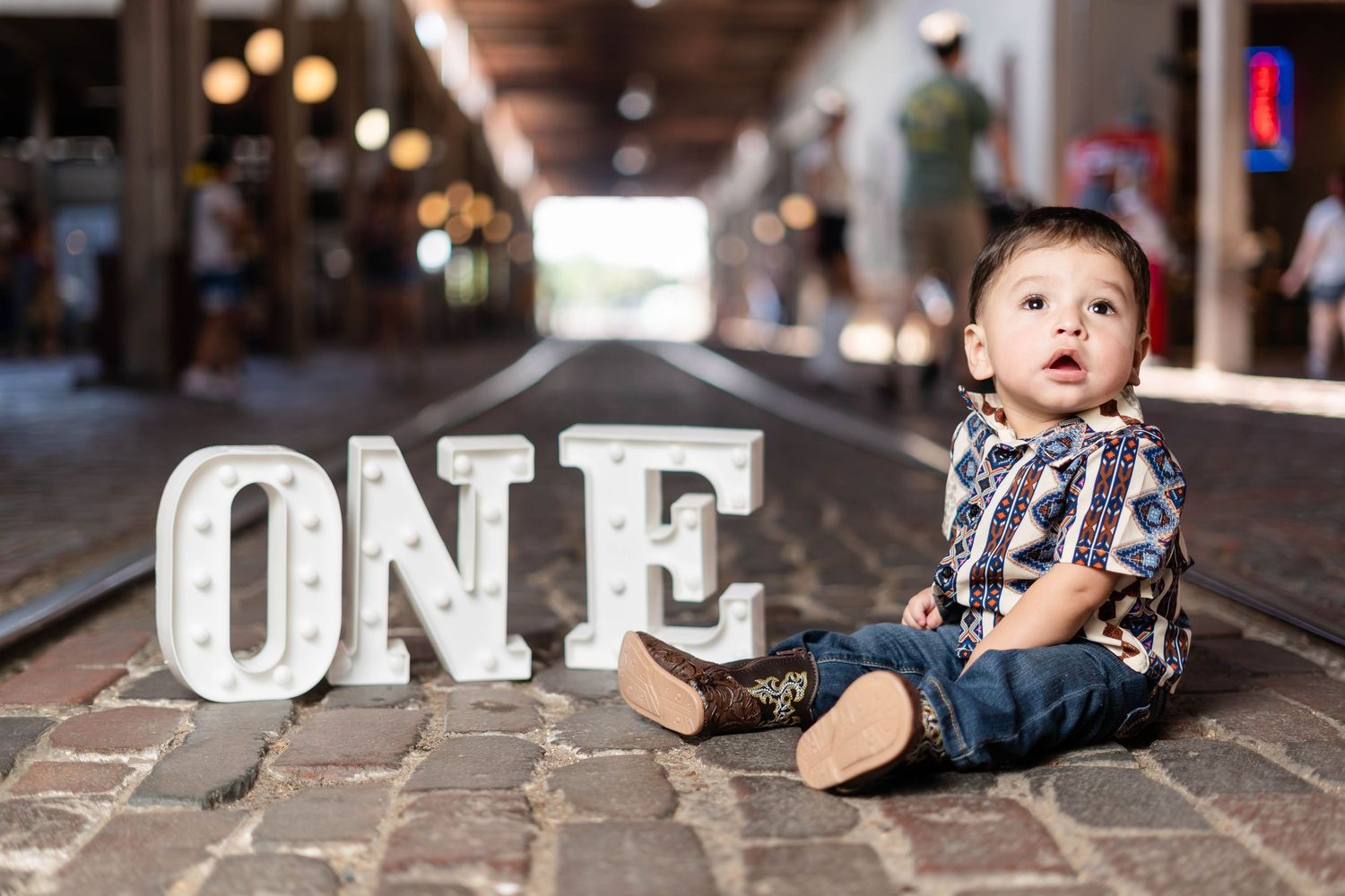 Baby boy in plaid shirt and jeans sitting on cobblestone path beside illuminated ONE marquee sign.
