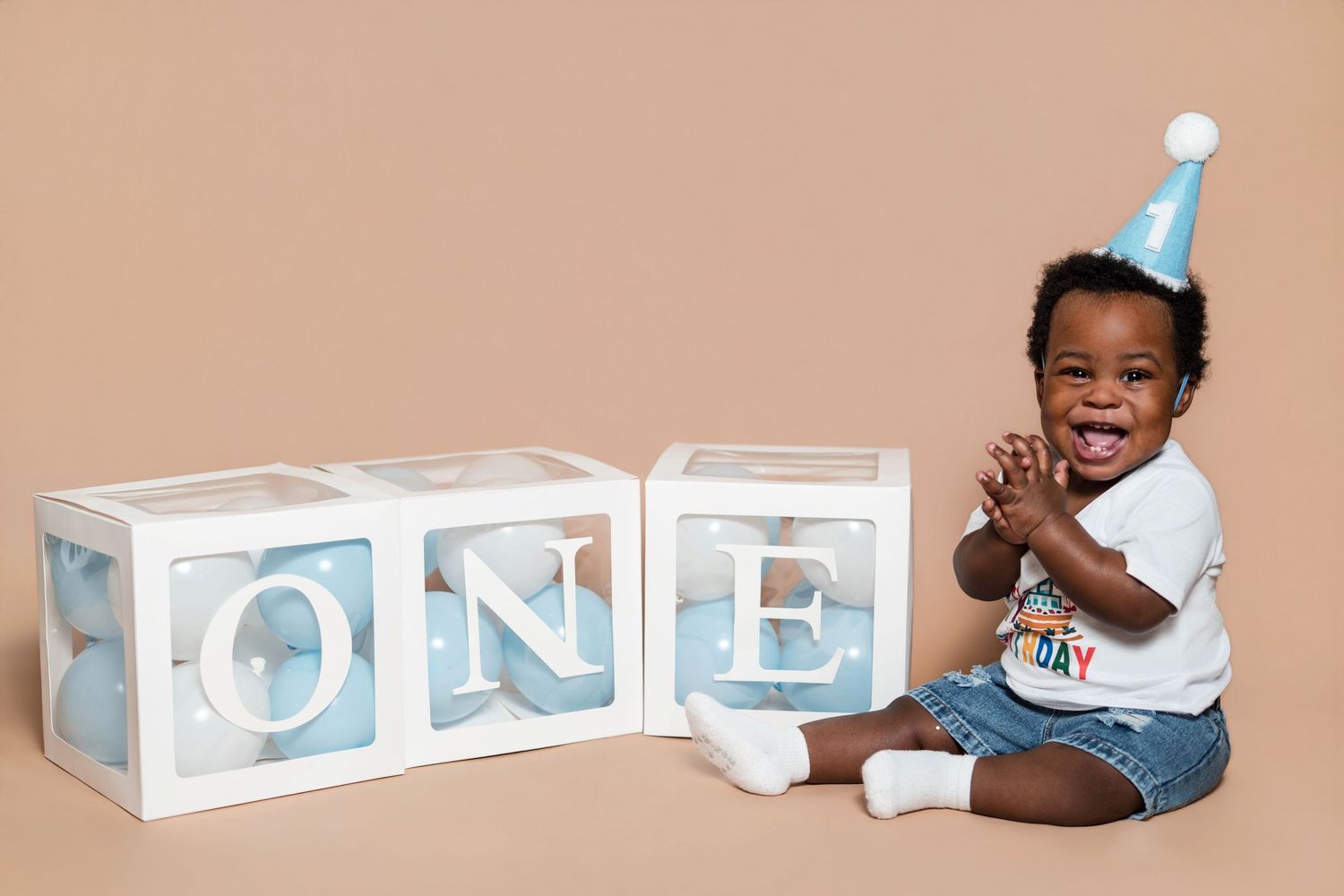 Smiling baby in birthday hat sits next to ONE letter blocks celebrating first birthday on beige background.
