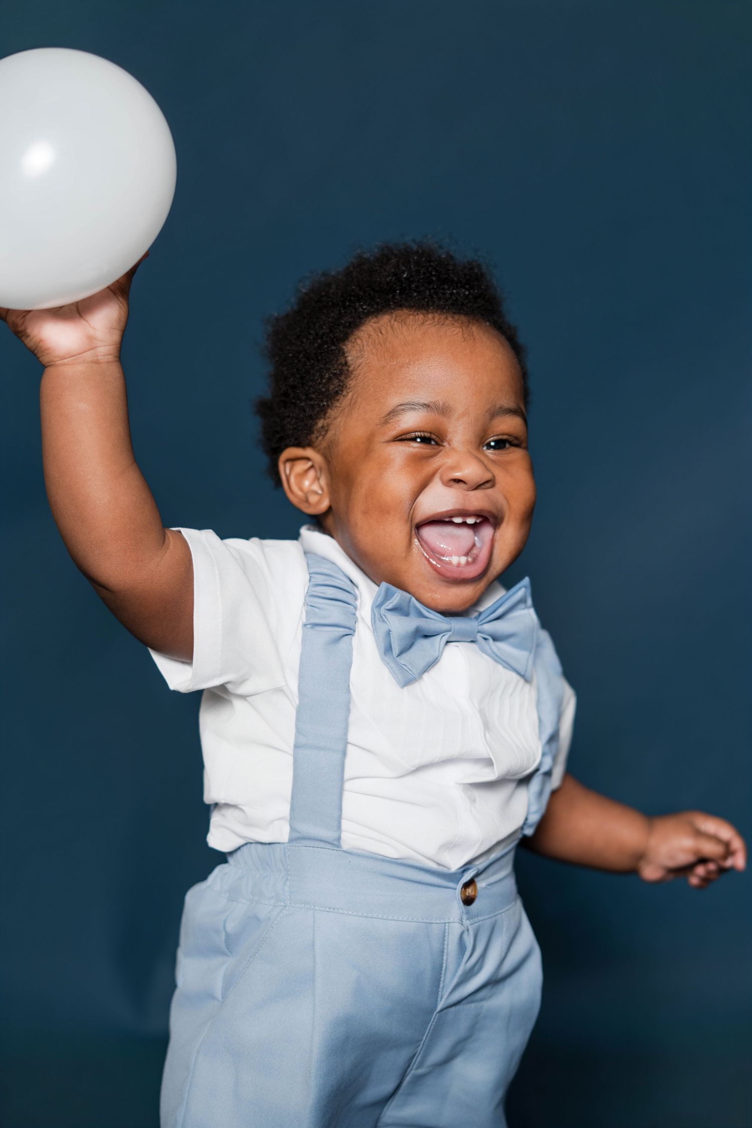 A joyful toddler boy in blue suspenders and bow tie holds a white balloon, laughing with mouth open wide.