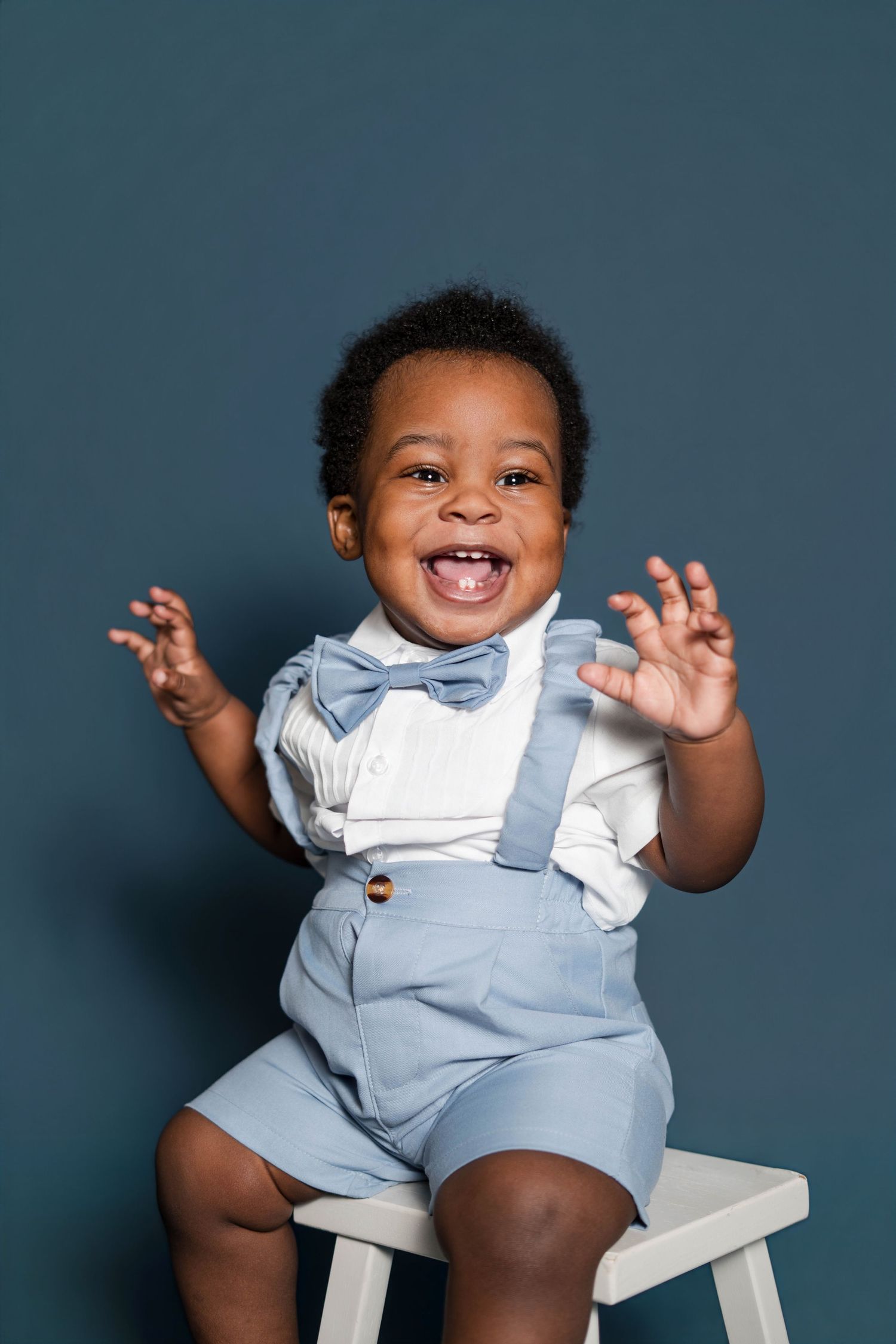 Adorable smiling Black toddler boy in light blue suspenders, shorts, and bow tie sitting on white stool against dark blue background.