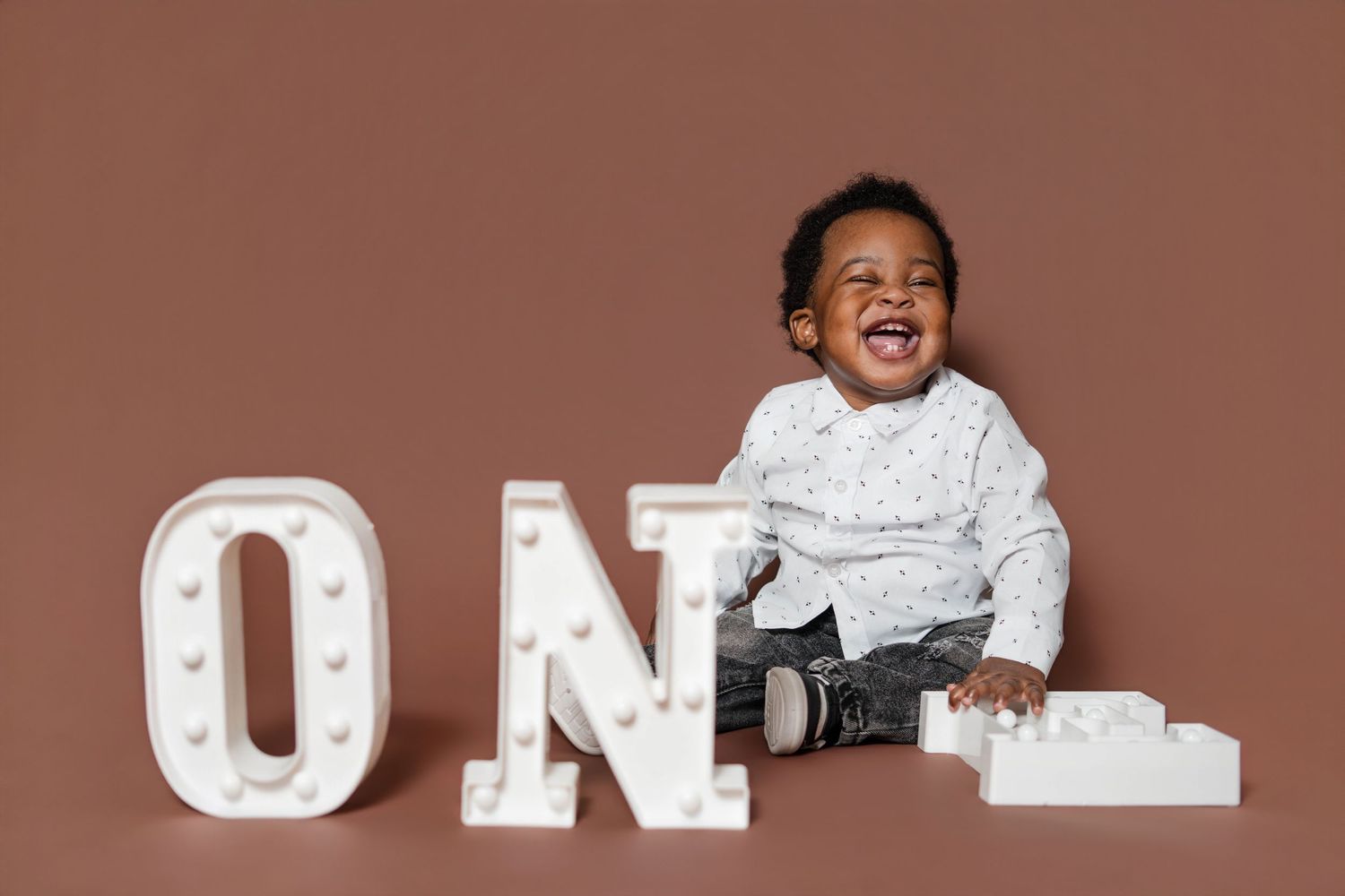 Laughing Black baby boy sitting next to illuminated ONE marquee letters on brown background for first birthday photo.