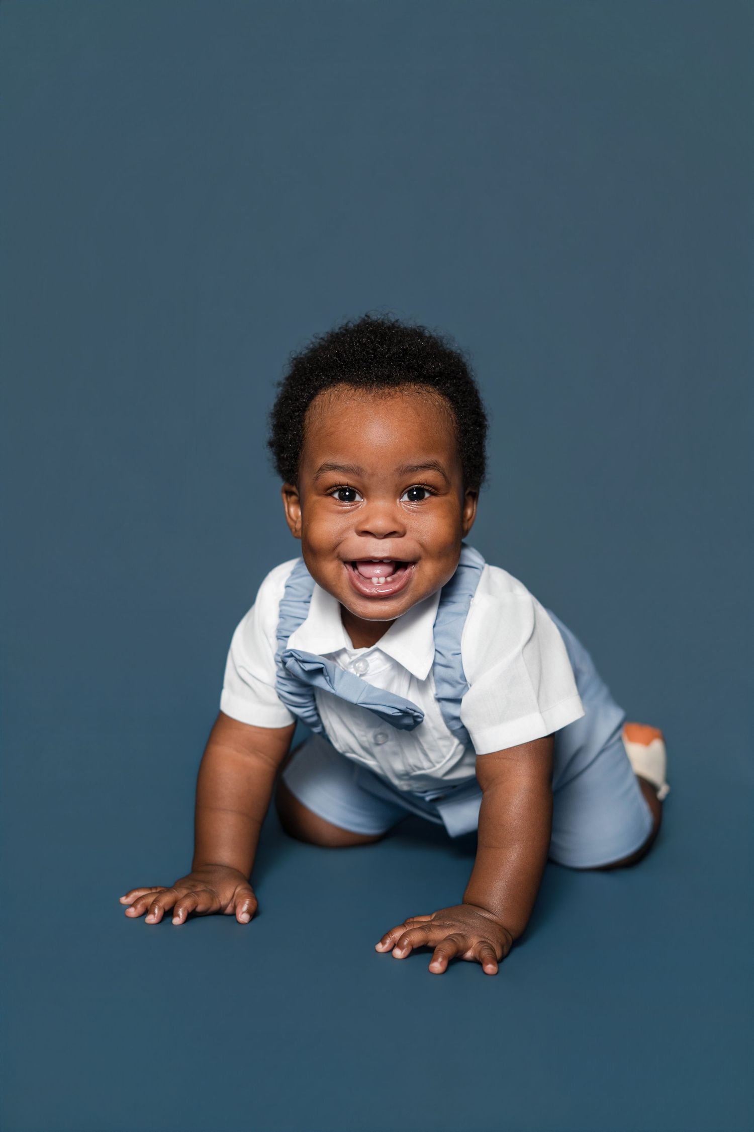 Smiling Black baby in white shirt and denim overalls crawling on a blue studio background.