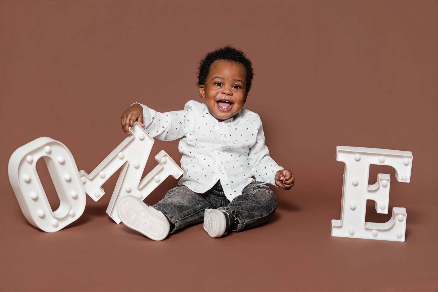 Smiling baby boy sitting on brown backdrop with white marquee letters spelling ONE for first birthday photo.