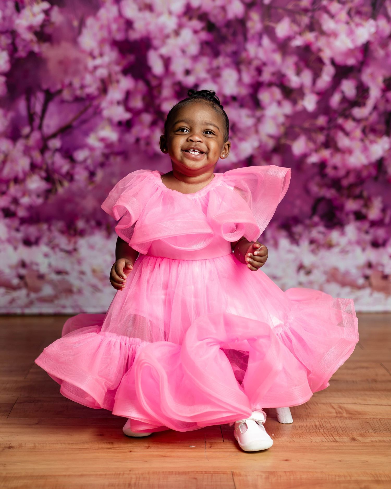 Joyful toddler girl twirling in a bright pink ruffle organza dress before floral backdrop.
