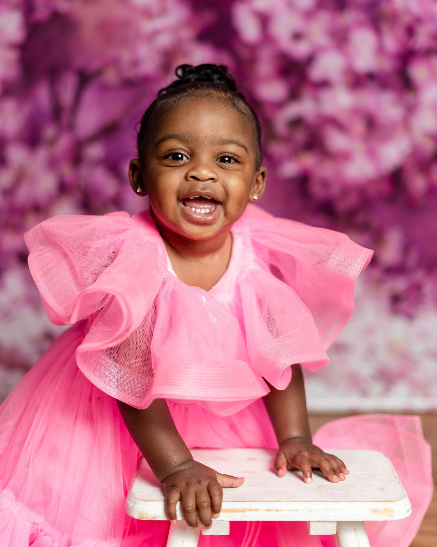 Smiling baby girl in pink tulle dress leaning on white stool with floral purple backdrop.