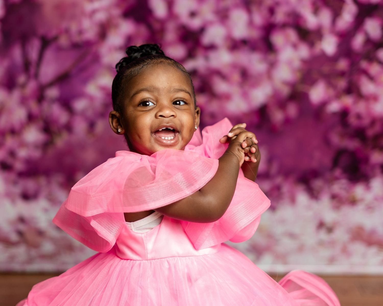 Happy baby girl in pink ruffled dress clapping hands in front of purple floral background.