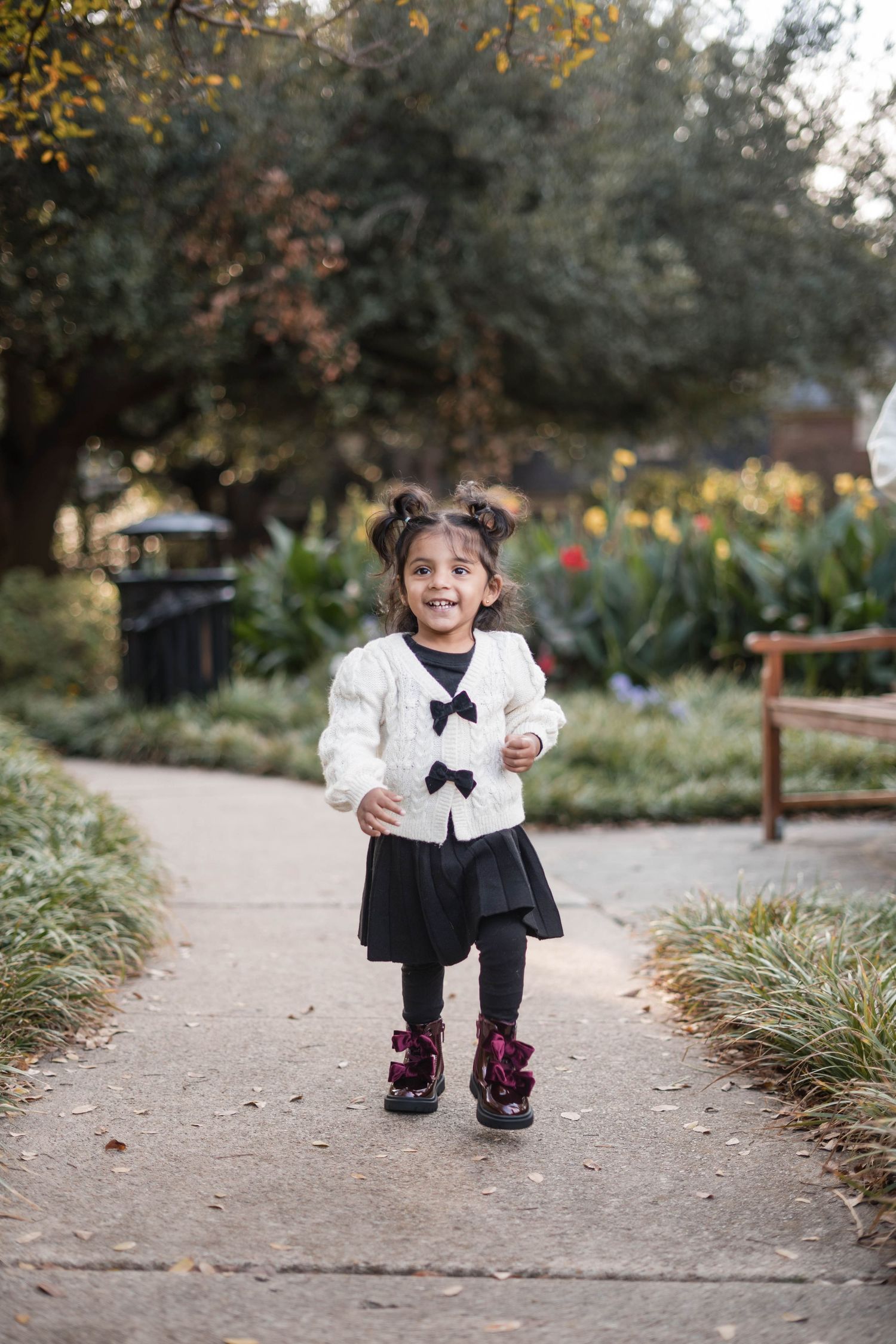 Cheerful toddler girl in white bow cardigan and black skirt running on garden pathway.