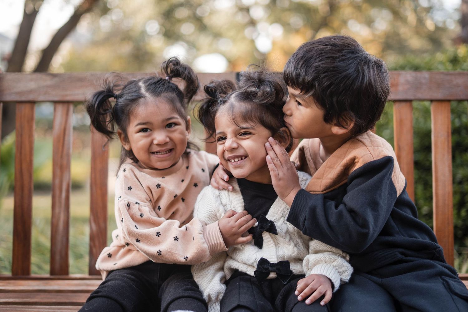 Three happy children sitting together on a wooden bench in a park, smiling and laughing.