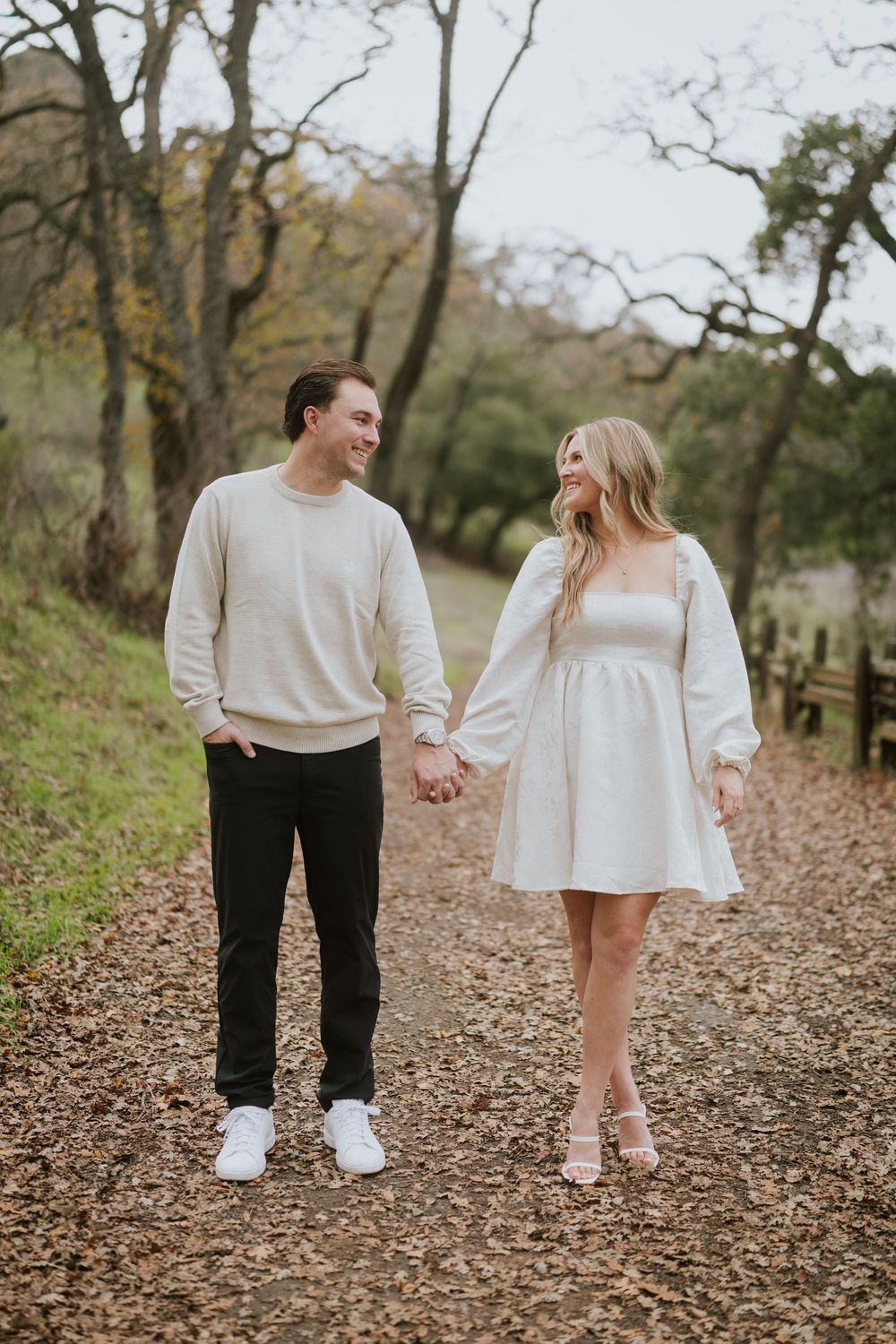 A couple walks hand in hand through a path covered in autumn leaves wearing coordinated cream-colored outfits.