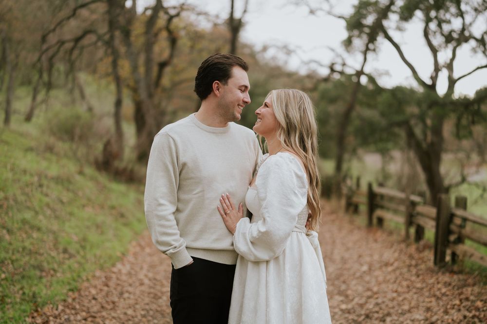 A romantic couple in white outfits shares tender moments along a wooded autumn path during a photo session.