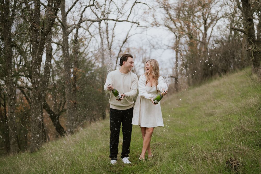 A couple shares a romantic moment together on a grassy hillside surrounded by bare trees in natural lighting.