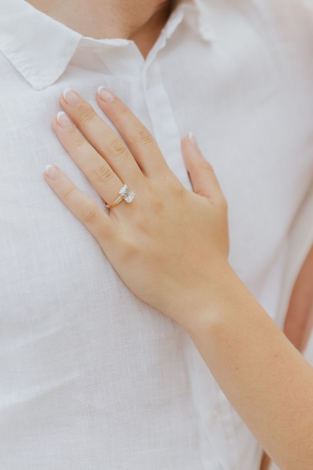 Close-up view of a hand wearing an elegant solitaire diamond engagement ring against a white fabric background.
