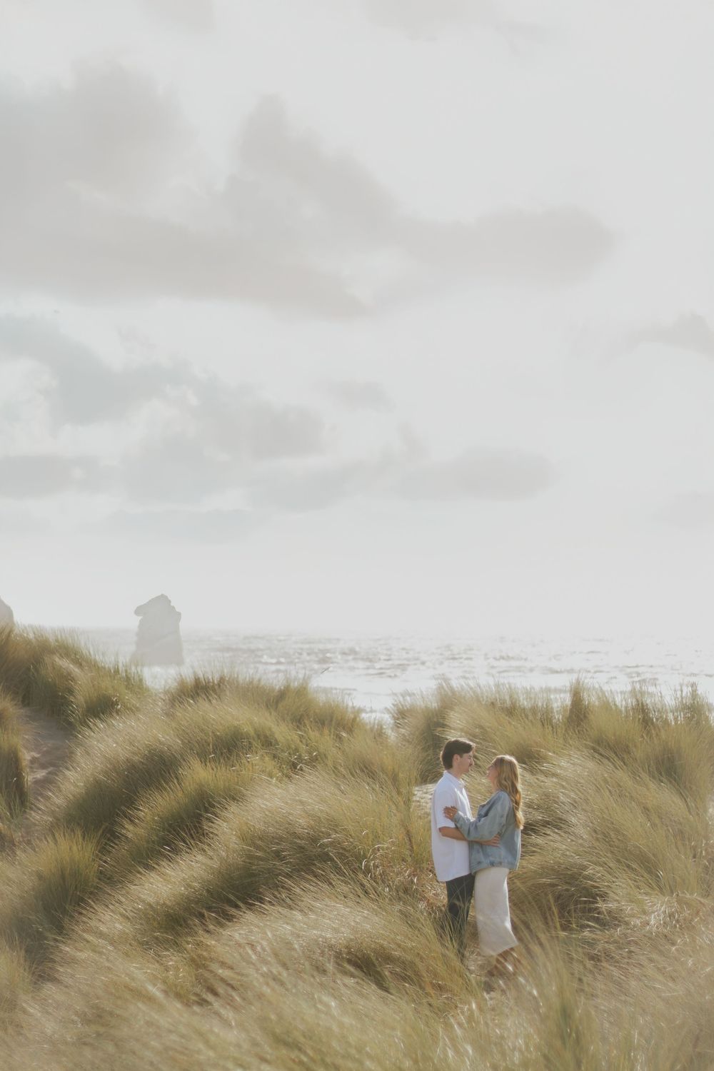 A couple stands together in tall beach grass as clouds roll across the coastal landscape at sunset.