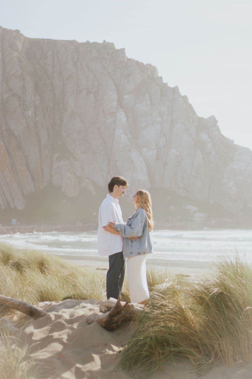 A couple embraces on a scenic beach with dramatic cliffs and coastal grasses in the background.