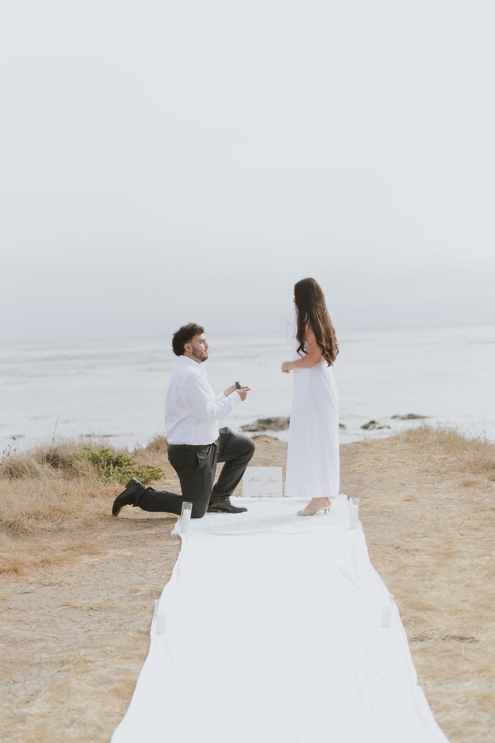 A romantic proposal scene on a coastal cliff with a white runner laid out, overlooking the ocean at sunset.
