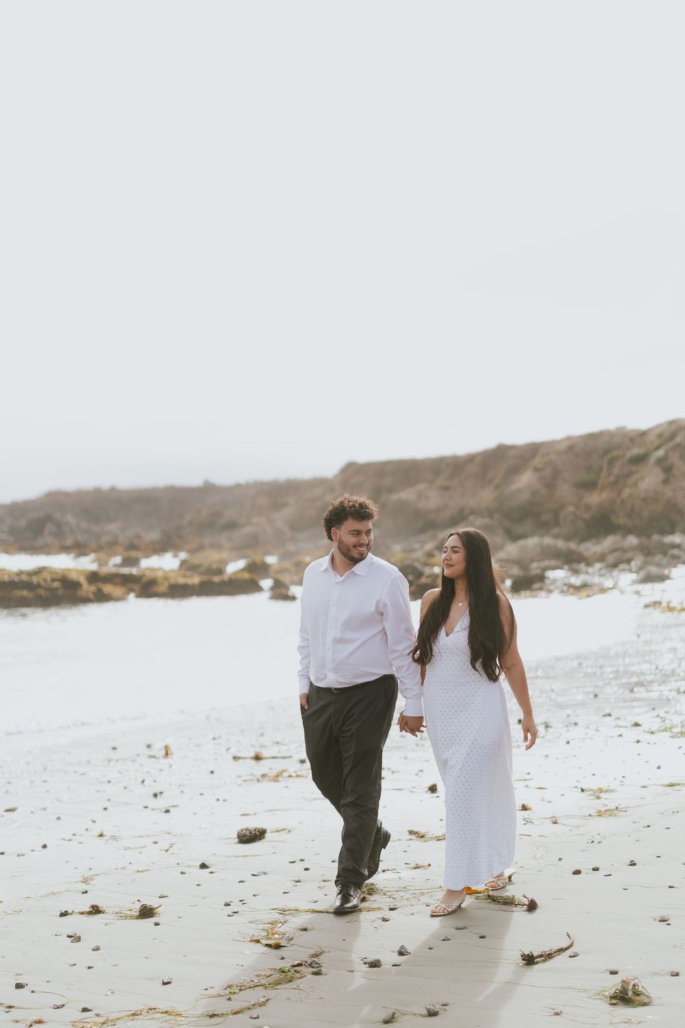 A couple in white outfits runs playfully along a misty beach shoreline.