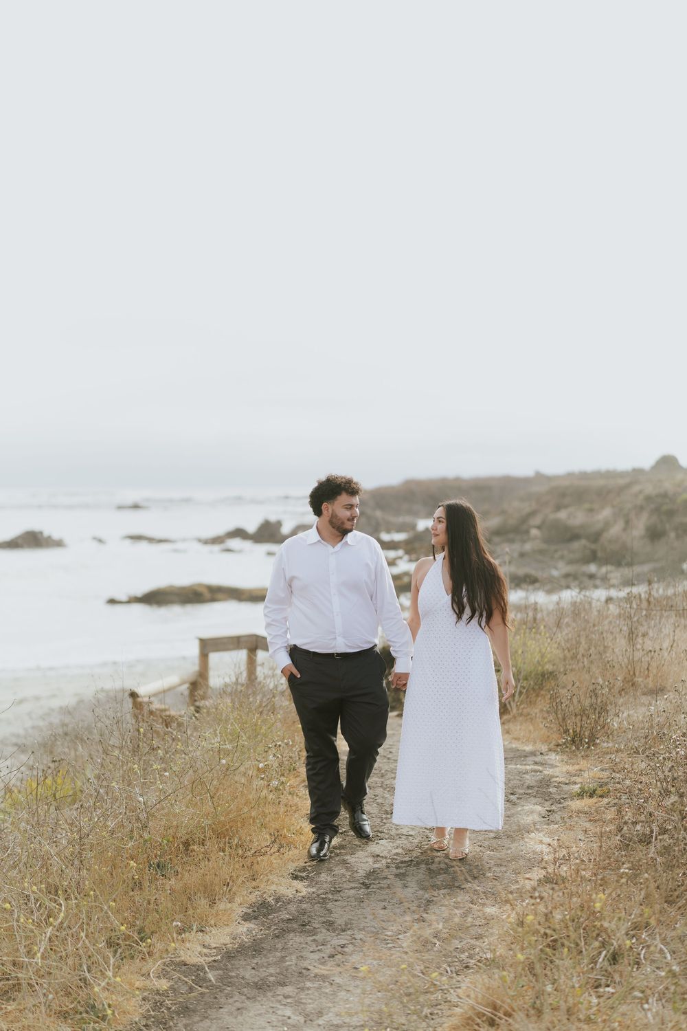 A couple walks along a coastal path through dry grass with ocean views on a cloudy day.