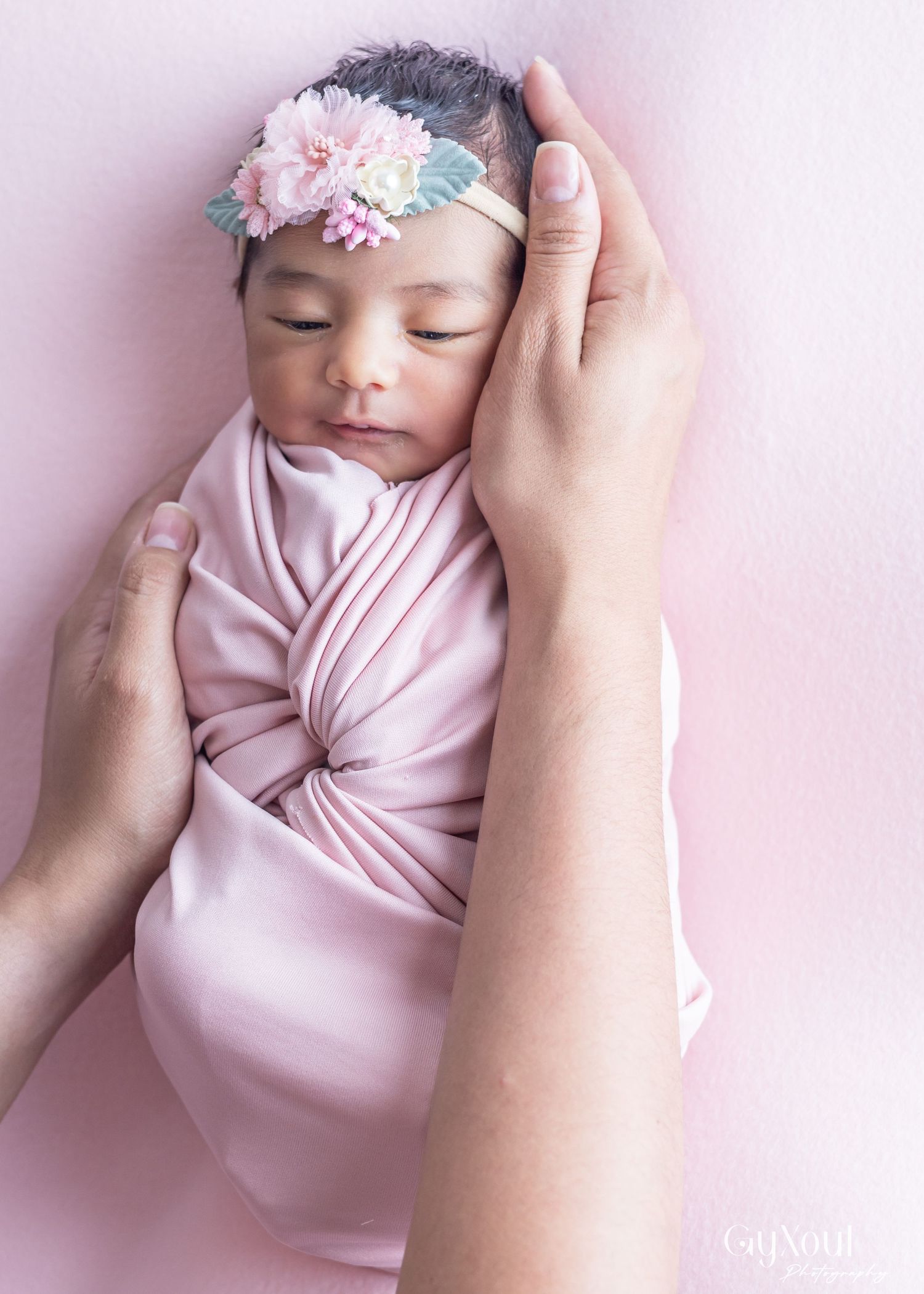Sleeping newborn wrapped in soft pink fabric with delicate floral headband on pink background.