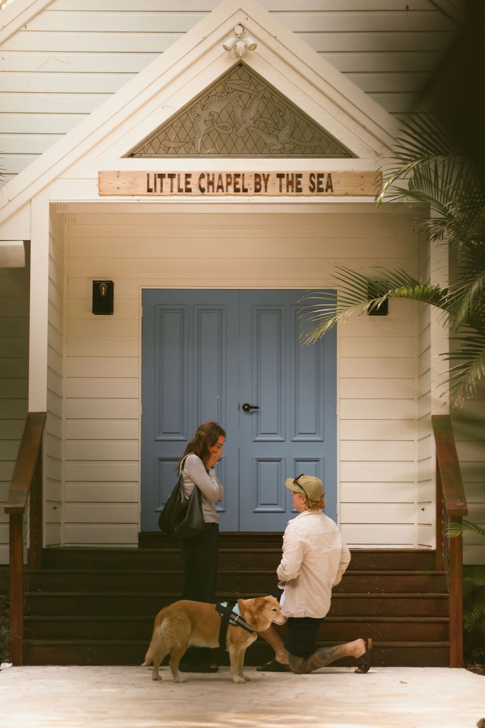 Two people and a dog stand at the entrance of the Little Chapel by the Sea, featuring a blue door and palm plants.