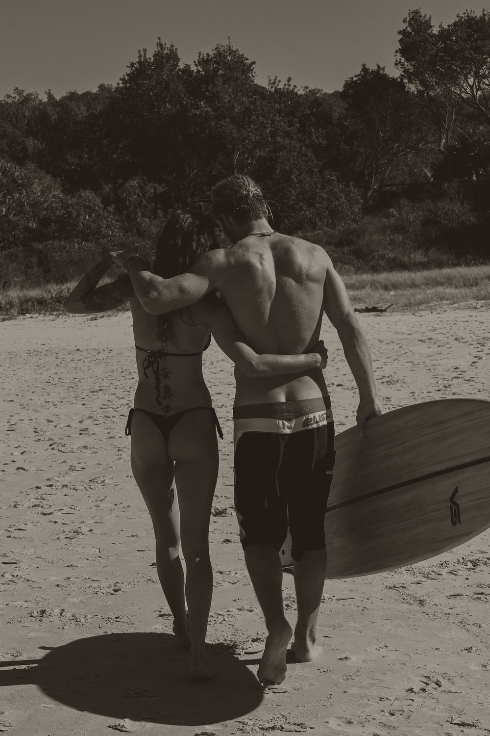 A group of surfers carrying their surfboards while walking along a sandy beach in black and white.