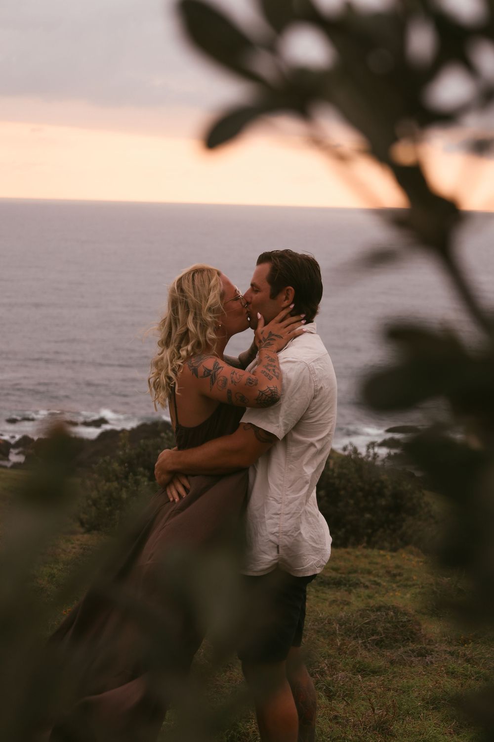 A romantic couple embraces on a coastal cliff at sunset with ocean waves crashing in the background.