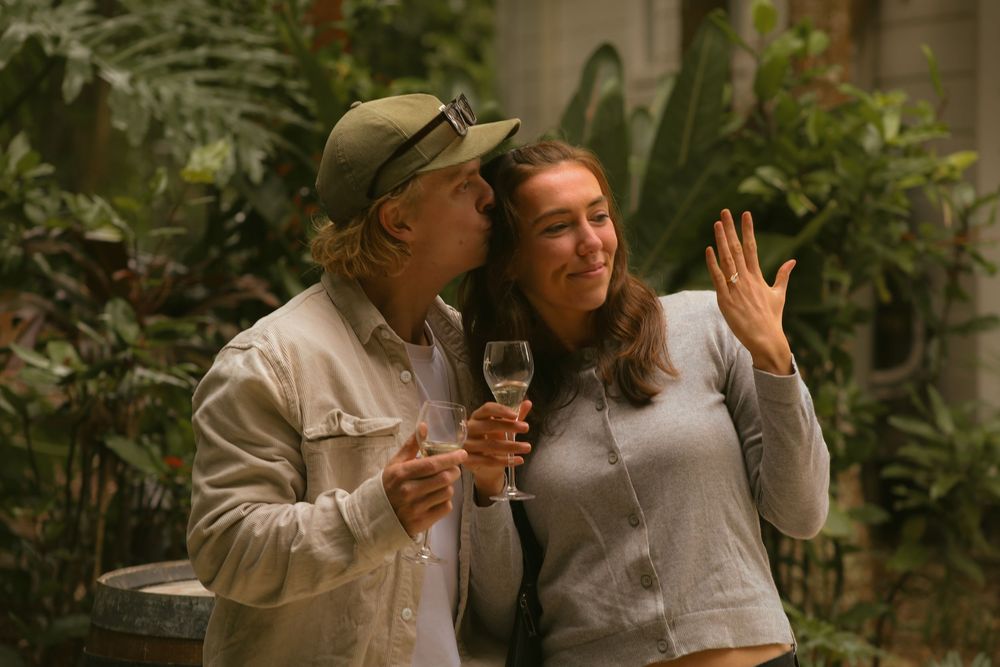 Two people in casual attire sit together at an outdoor table while sharing drinks and conversation in a garden setting.