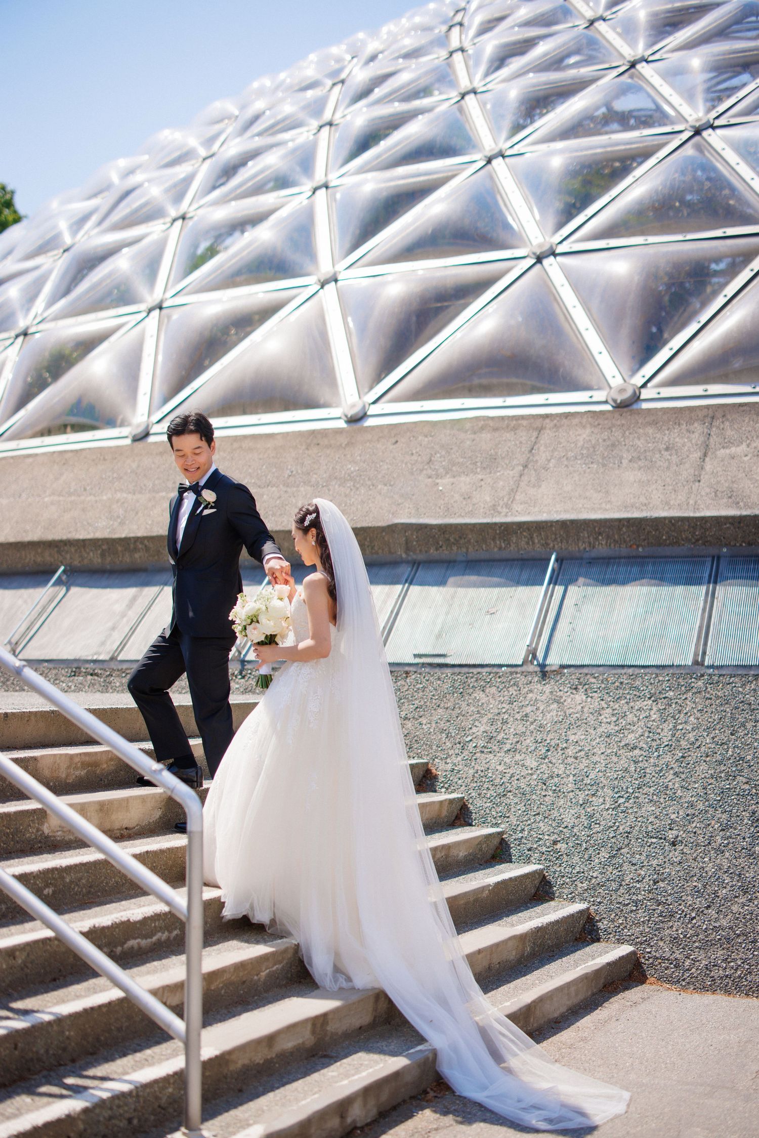 A wedding couple poses on concrete steps against a modern geometric domed structure with triangular glass panels.