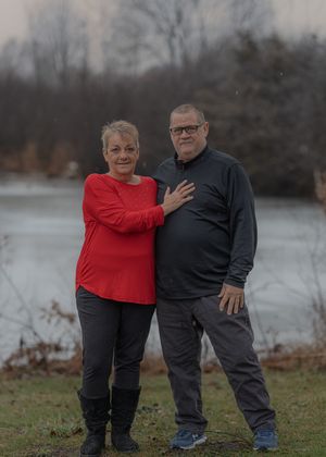 Couple in red and black casual wear poses together by a lake on an overcast day.