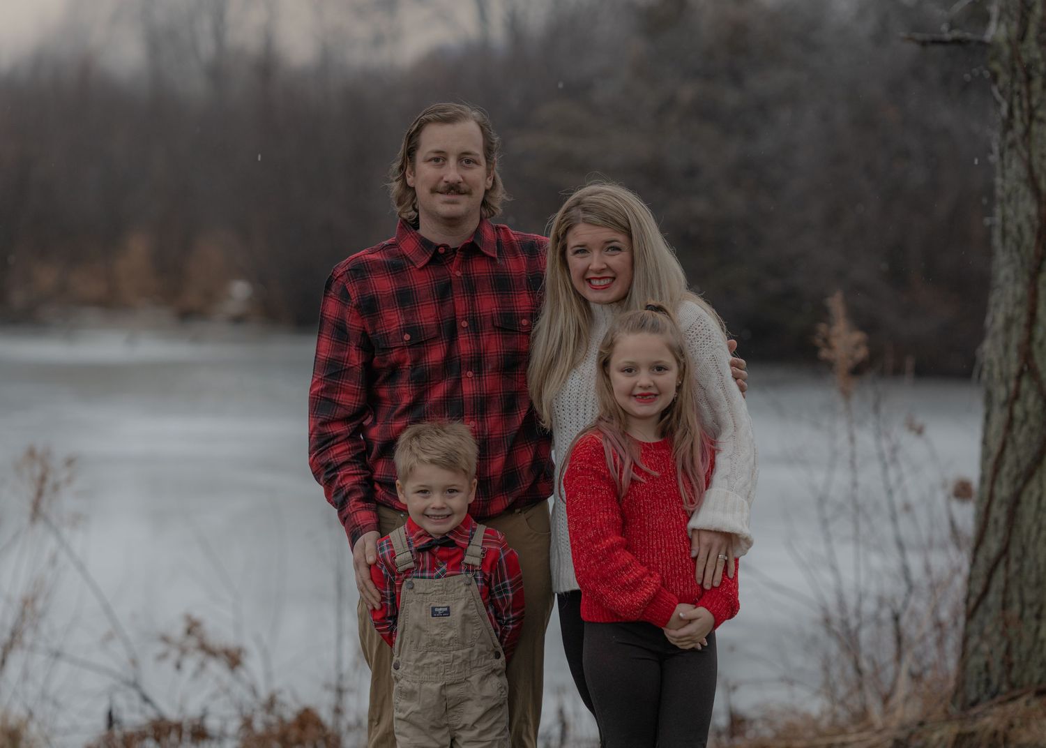 A family of four poses together by a lake wearing coordinated red and plaid outfits.