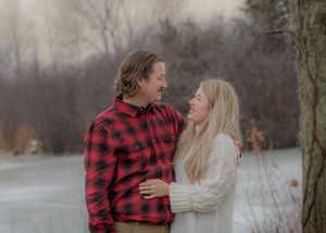 Couple embraces while wearing buffalo plaid and white sweater against winter lake backdrop.