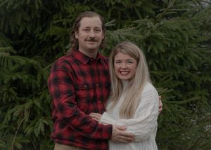 A couple embraces in front of evergreen trees wearing plaid and white winter clothing.