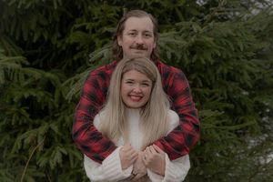 A couple shares a tender moment in front of pine trees wearing coordinated winter outfits.