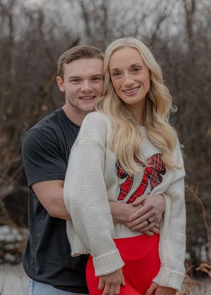 Couple in casual wear embraces while smiling at camera against winter forest background.