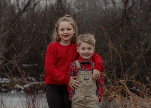 Two siblings pose together outdoors wearing coordinated red winter outfits.