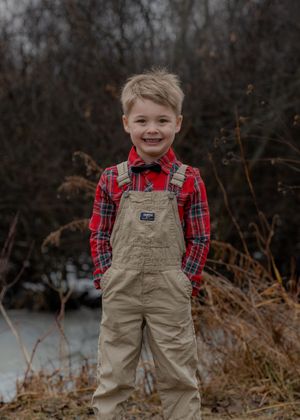 A child in red plaid shirt and tan overalls smiles brightly outdoors.