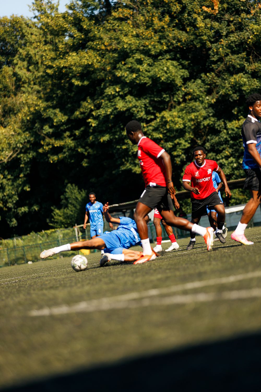 Soccer players in red and blue jerseys engage in a sliding tackle during an outdoor match on a grassy field.
