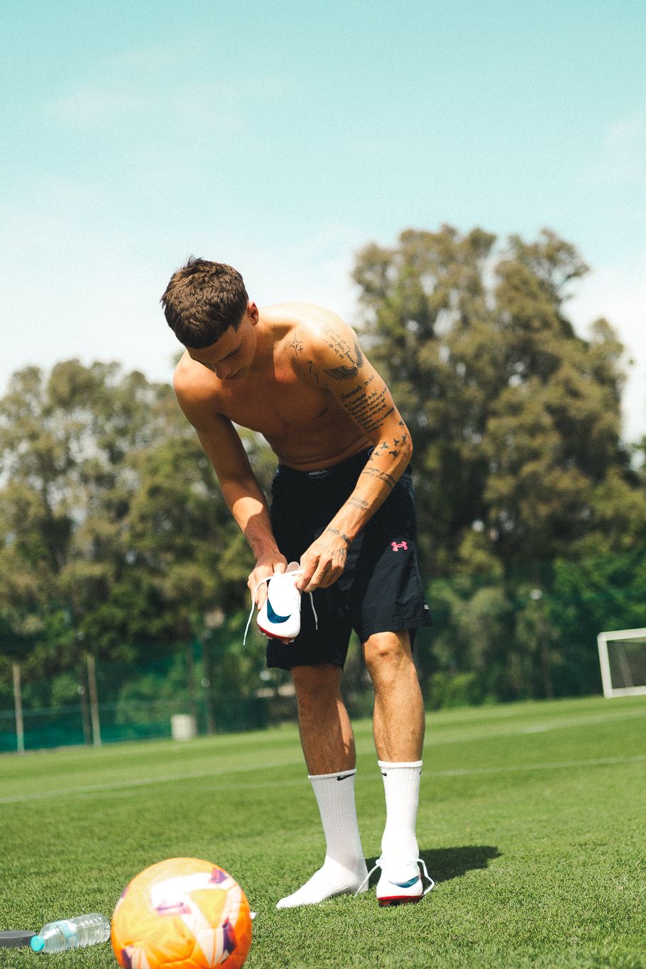 Shirtless tattooed footballer adjusting cleats on sunny green pitch with soccer ball nearby.