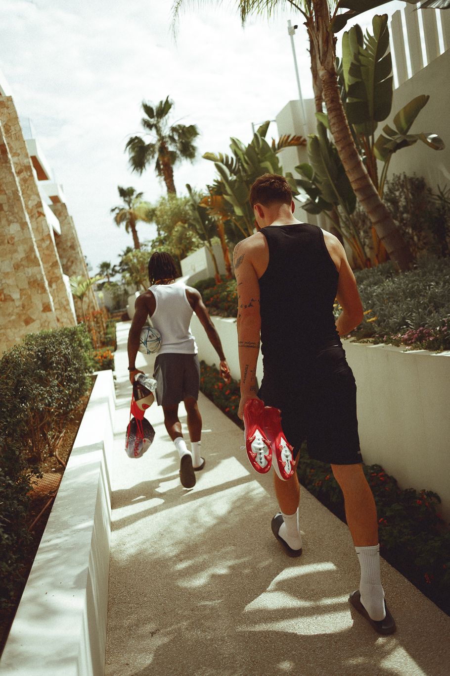 Two men in athletic wear carry red sneakers while walking along a sunny palm-lined path near white buildings.