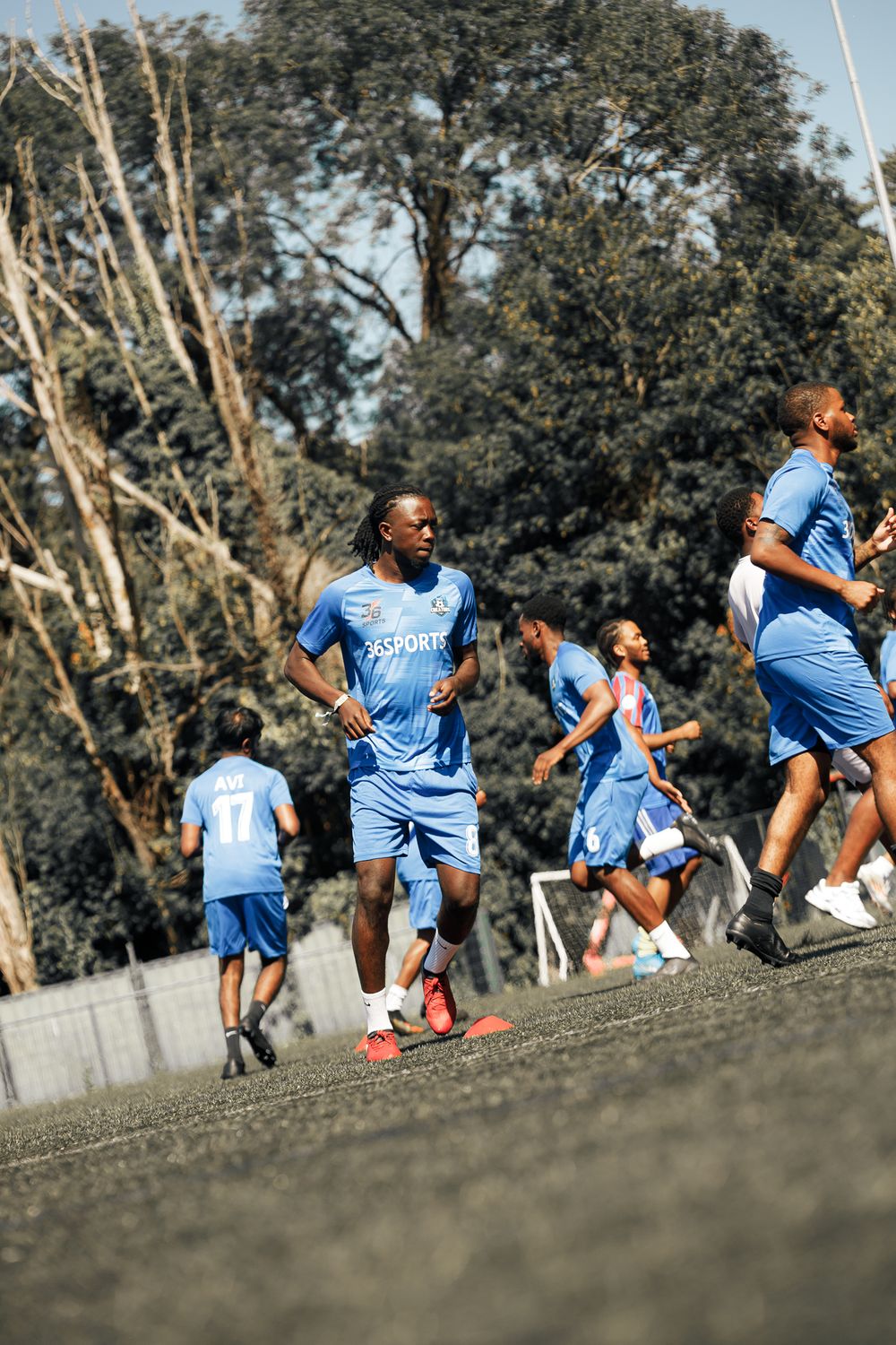 Athletes in light blue uniforms perform dynamic warm-up exercises on a grassy field with trees in the background.