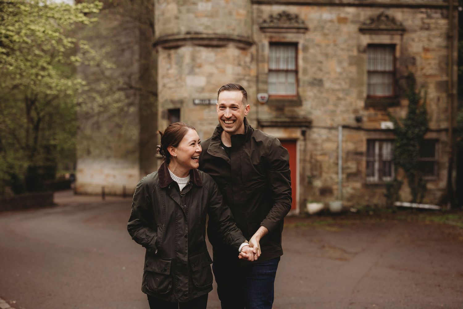 A smiling couple holds hands while walking in front of a historic stone building surrounded by lush green trees.