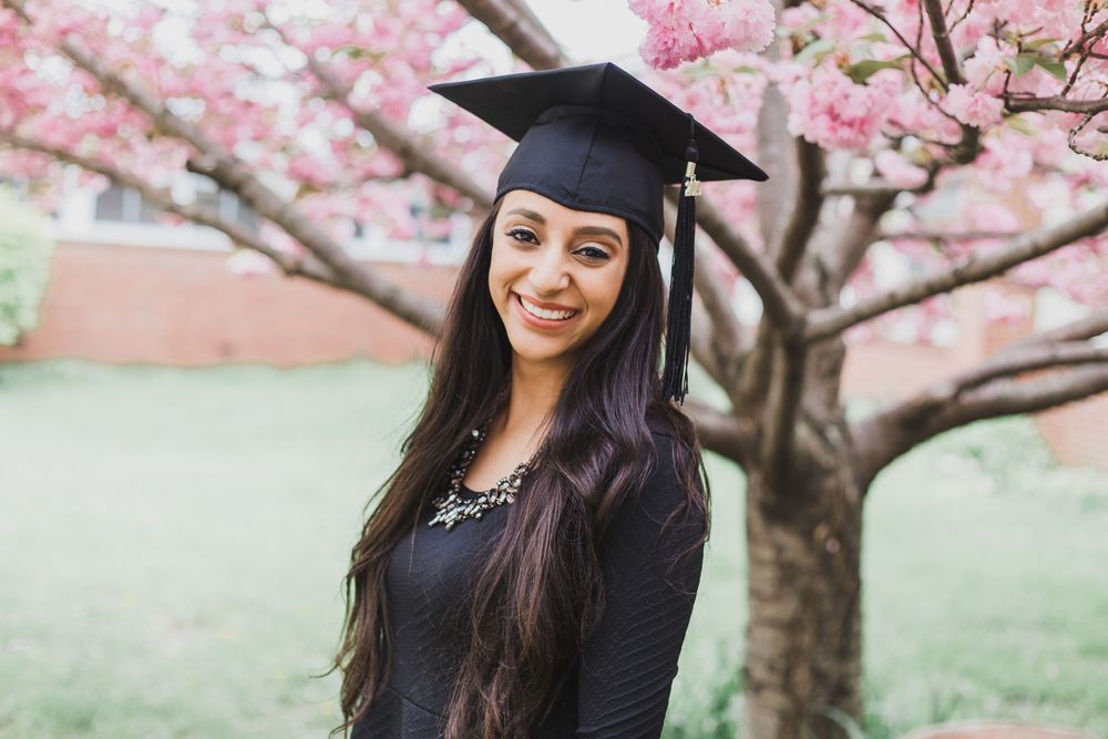 Graduate wearing mortarboard poses beneath pink cherry blossoms during spring graduation photoshoot.
