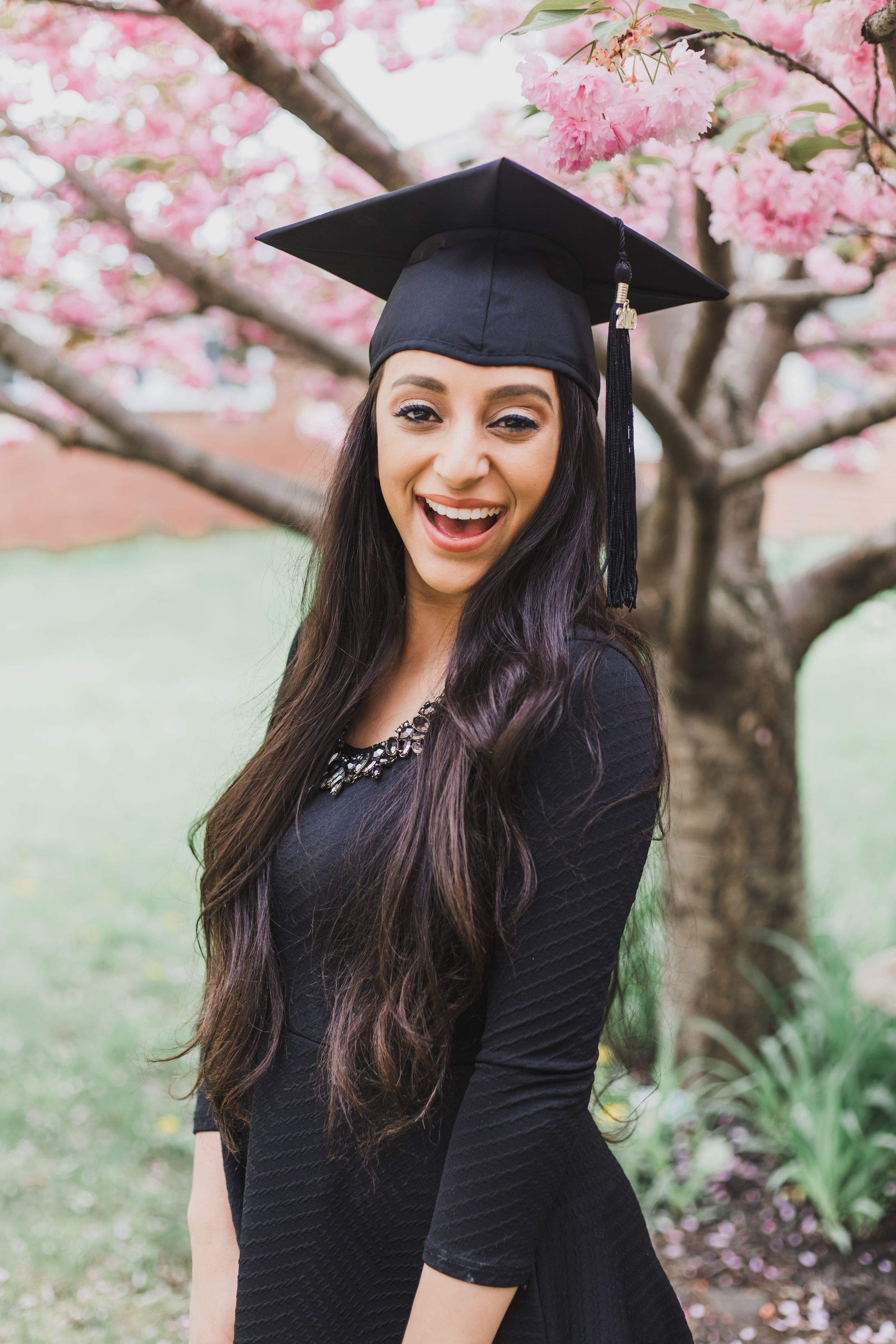 Graduate in black dress and cap smiling under blooming cherry blossom trees during spring photo session.