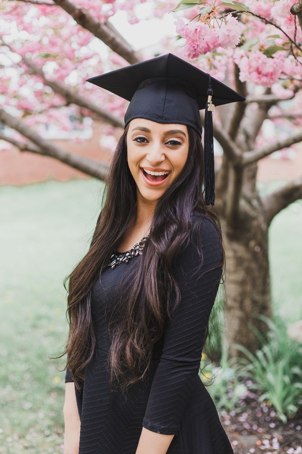 Graduate in black dress and cap smiling under blooming cherry blossom trees during spring photo session.