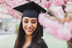 Graduate in cap and gown smiles among blooming pink cherry blossoms during spring graduation photo session.